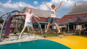 Dos niños saltando sobre un trampolín frente a un edificio con techo rojo y un área de patio.