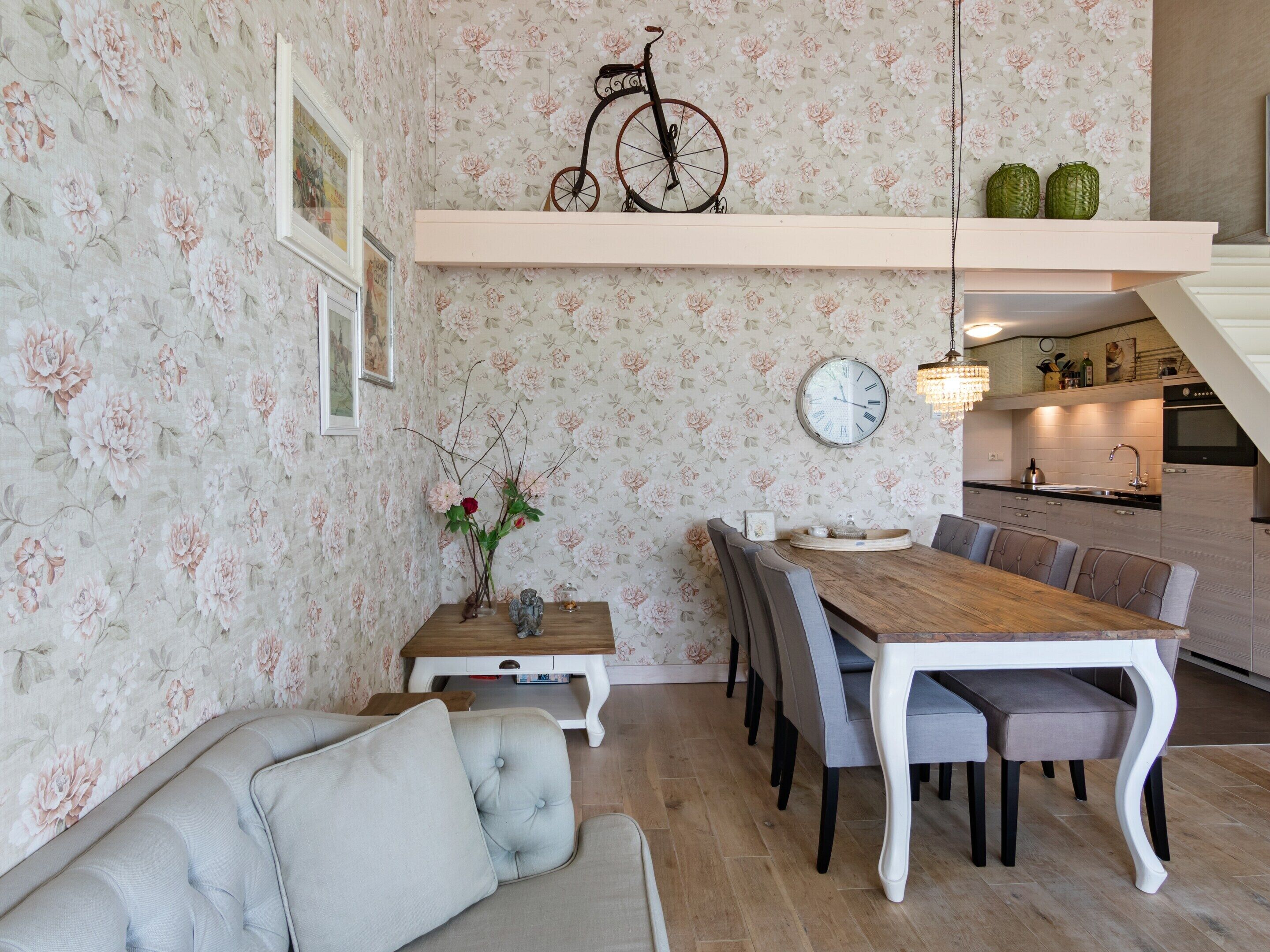 A dining area with a wooden table and chairs, a floral wallpaper, a bicycle decoration, and a kitchen in the background.