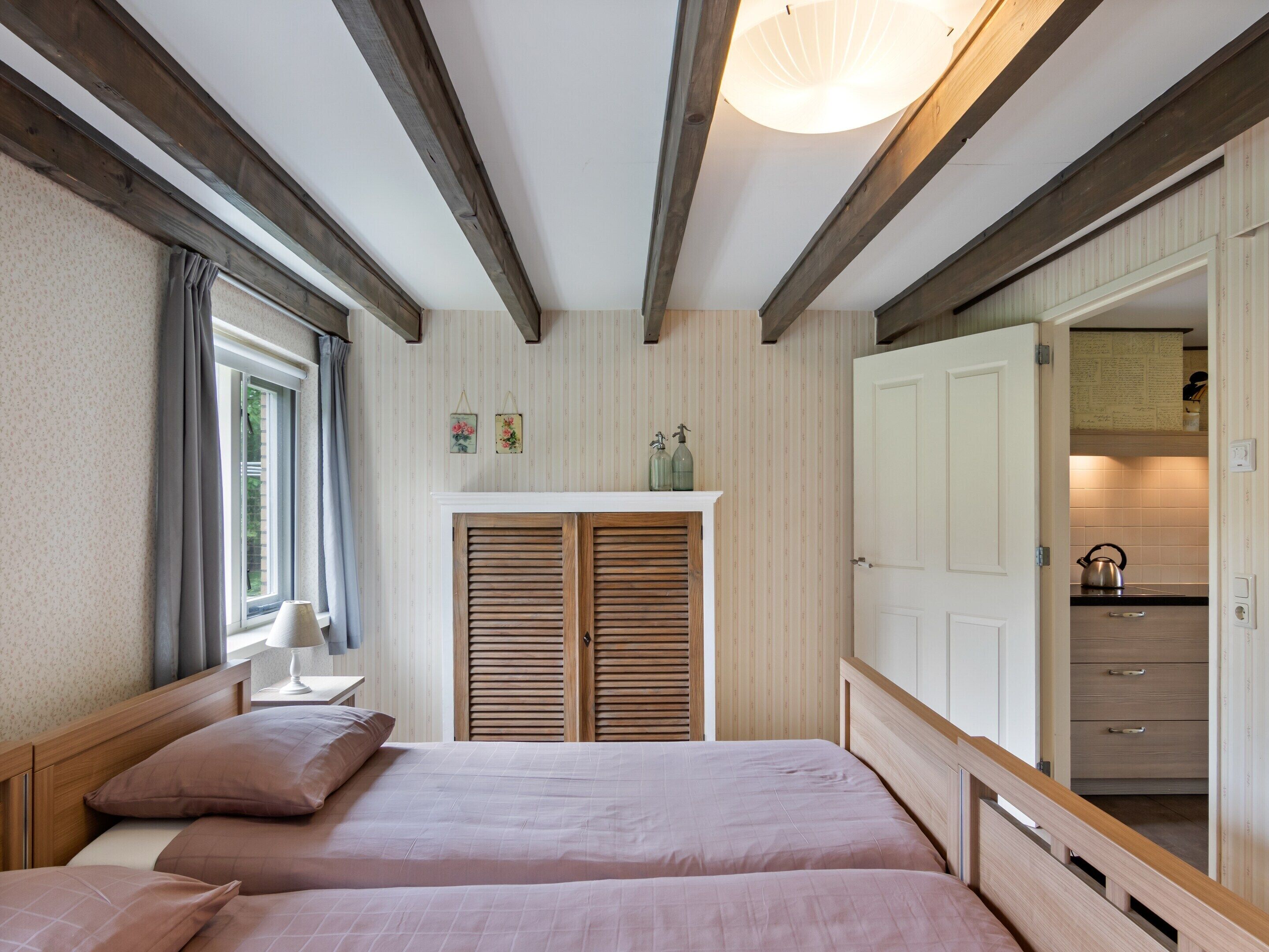 A bedroom with two beds, a window with curtains, a bedside table, a shelf with decorative items, and a view into the kitchen.