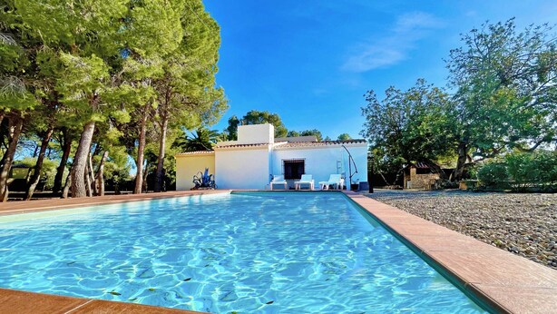 Une piscine à l’eau d’un bleu cristallin, entourée d’arbres et bordée d’un bâtiment dont le toit est recouvert de tuiles.