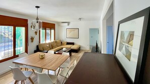 Un salon moderne avec une table à manger en bois, des chaises blanches, un canapé beige et un grand tableau encadré au mur.