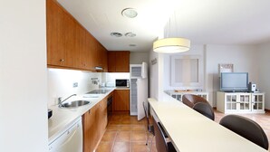 A compact kitchen with wooden cabinets, a built-in microwave, a refrigerator, and a dining area with a TV and a small cabinet. - ORQUIDEA LES OLIVERES (El Perelló)