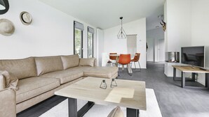 Una sala moderna con un sofá seccional color beige, una mesa de centro de madera y un comedor con sillas naranjas.