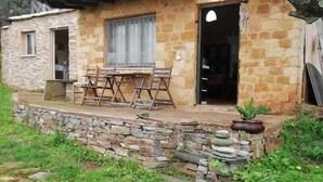 A stone house with a tiled roof, a small patio with a table and chairs, and a garden area with a hose.