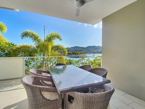 Un balcon doté d’une table à plateau en verre et de chaises en osier, offrant une vue sur une étendue d’eau et des collines.