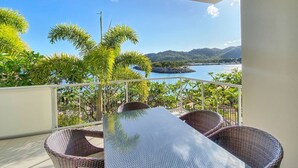 A balcony with wicker furniture, a glass table, and a view of a coastal landscape. - 1 Bright Point Apartment 2204 (Nelly Bay)