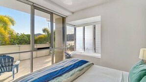A bedroom with a large bed, a window with blinds, a balcony with a chair, and a view of greenery. - 1 Bright Point Apartment 2204 (Nelly Bay)