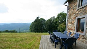 A wooden deck with a table and chairs, overlooking a grassy area and a stone building with a window.