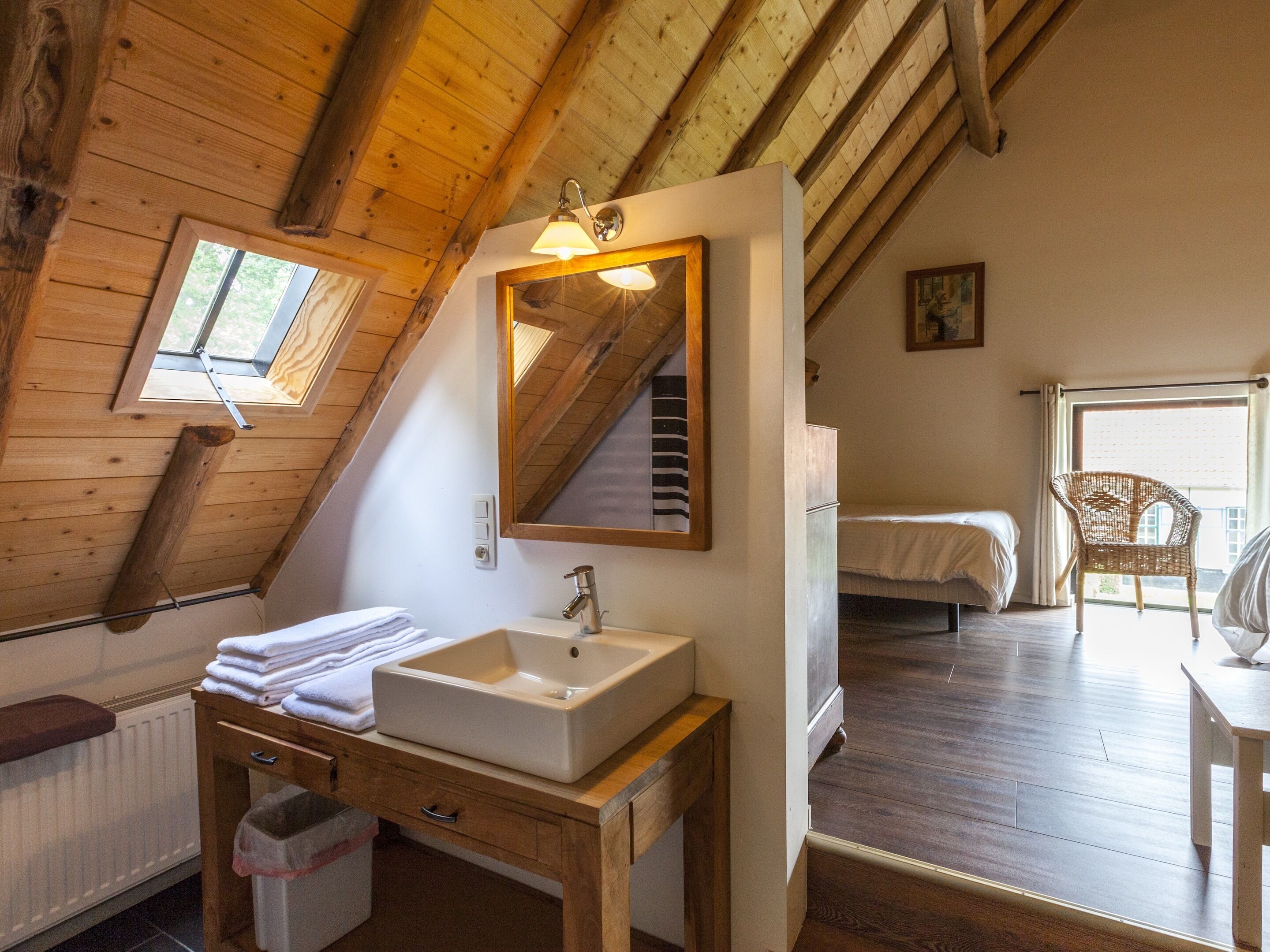 Um quarto de sótão aconchegante, com teto de madeira, um espelho grande, uma pia branca, uma penteadeira de madeira, uma cama e uma cadeira de vime.