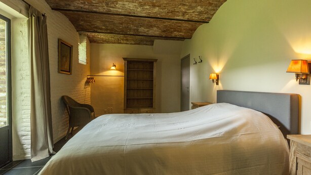 A bedroom with a large bed, a wooden headboard, a nightstand, a wall-mounted lamp, and a window with curtains.