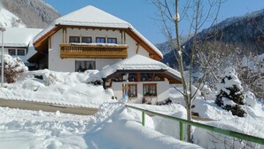 Chalé de dois andares com telhado coberto de neve e varanda, em meio a uma paisagem nevada e montanhas.