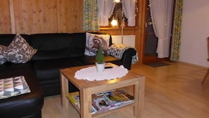 Uma sala de estar acolhedora com um sofá preto, uma mesa de centro de madeira com revistas, uma pequena planta em vaso e uma janela com cortinas.