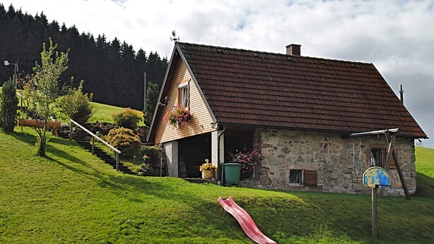 A rustic stone house with a red-tiled roof, a wooden staircase, and a red slide on a grassy hill. - Ferienhaus in Brigach (Sankt Georgen im Schwarzwald)