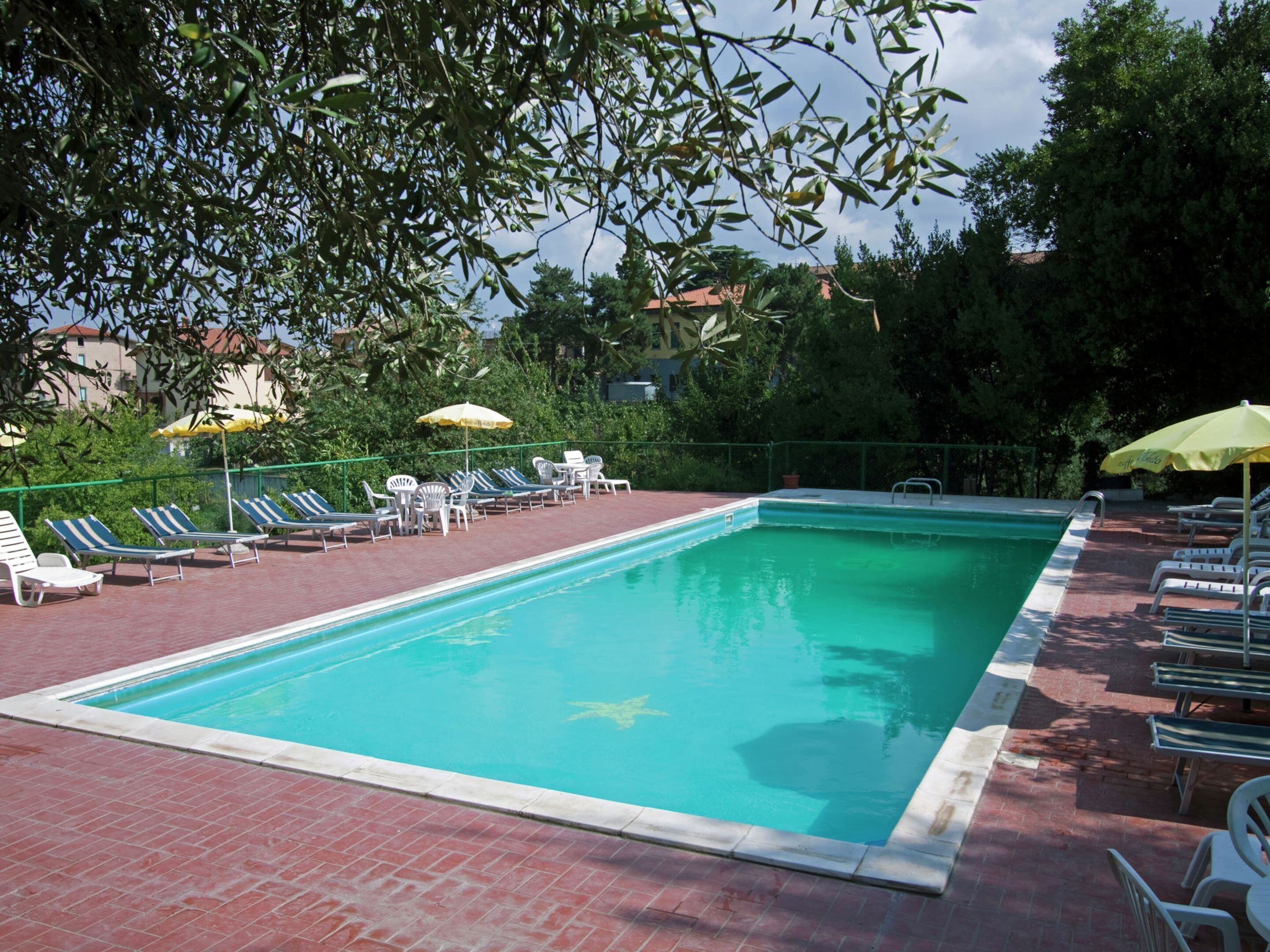 A rectangular swimming pool with a tiled edge, surrounded by a brick deck. There are lounge chairs and umbrellas along the poolside.