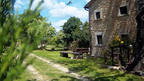 Un edificio de piedra con una mesa de picnic y bancos en un césped, rodeado de árboles y un cielo despejado.