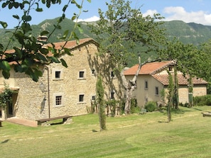 A stone building with multiple windows and a red-tiled roof, surrounded by a grassy area and trees.