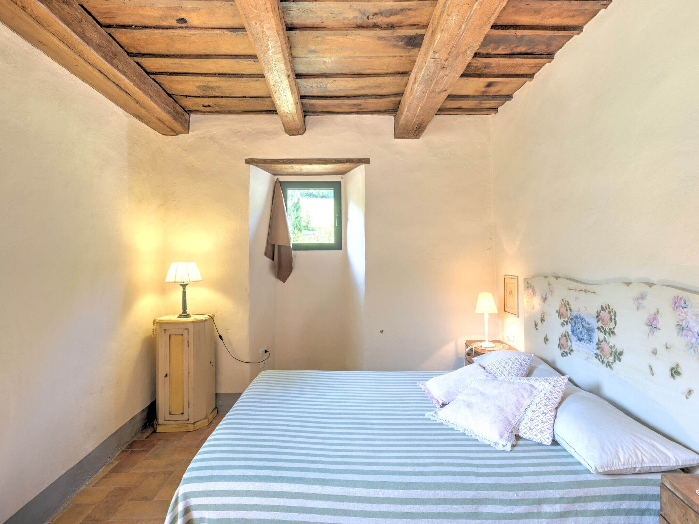 Un dormitorio con techo de madera, una ventana pequeña, una cama con ropa de cama a rayas, dos lámparas de noche y un cabecero floral.