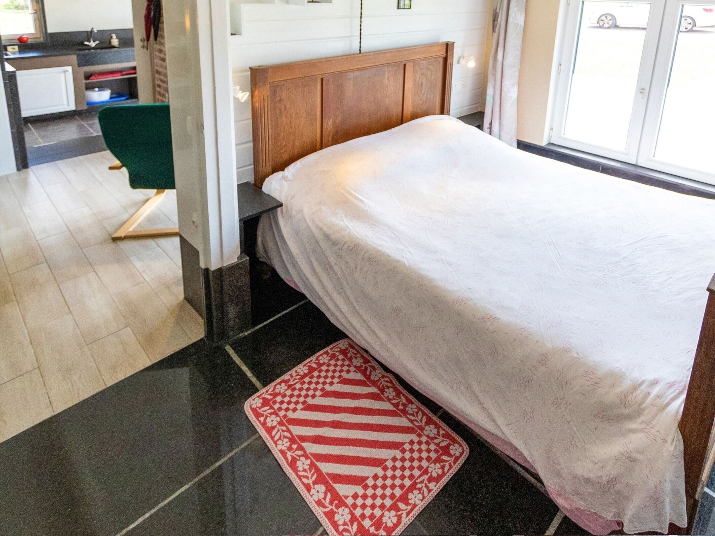 A neatly made bed with a white bedspread, a red and white patterned rug, and a wooden headboard.