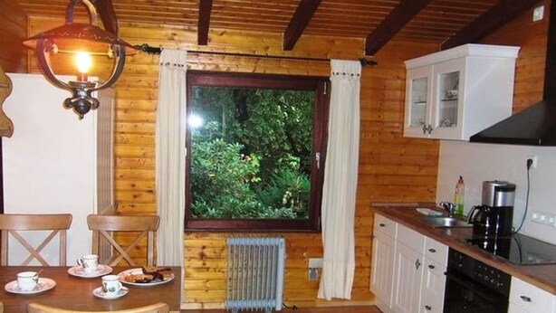 A cozy cabin-style room with a wooden interior, a dining table with chairs, a window with curtains, a radiator, and a kitchenette with cabinets and appliances. - Dog-friendly Holiday Home in the Knull (Neuenstein)