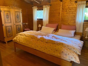 A wooden cabin bedroom with a bed, two chairs, a wardrobe, and a window with curtains. - Dog-friendly Holiday Home in the Knull (Neuenstein)