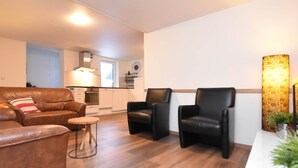 A modern living room with a brown leather sofa, two black leather armchairs, and a small round table. A kitchen is visible in the background with white cabinets and a stainless steel oven.