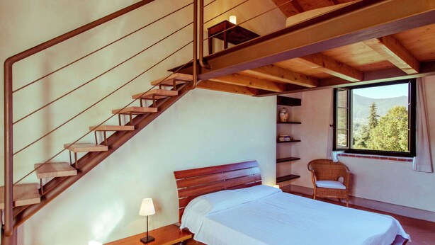 A bedroom with a wooden staircase, a bed with a white bedspread, a chair, and a window with a view of trees. - Belvilla by OYO Bilo Grande Della Cascina (Salò)