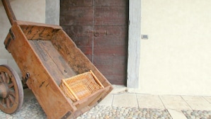 A wooden cart with a wicker basket, positioned in front of a large, dark wooden door with a stone frame, set against a plain wall. - Belvilla by OYO Bilo Grande Della Cascina (Salò)