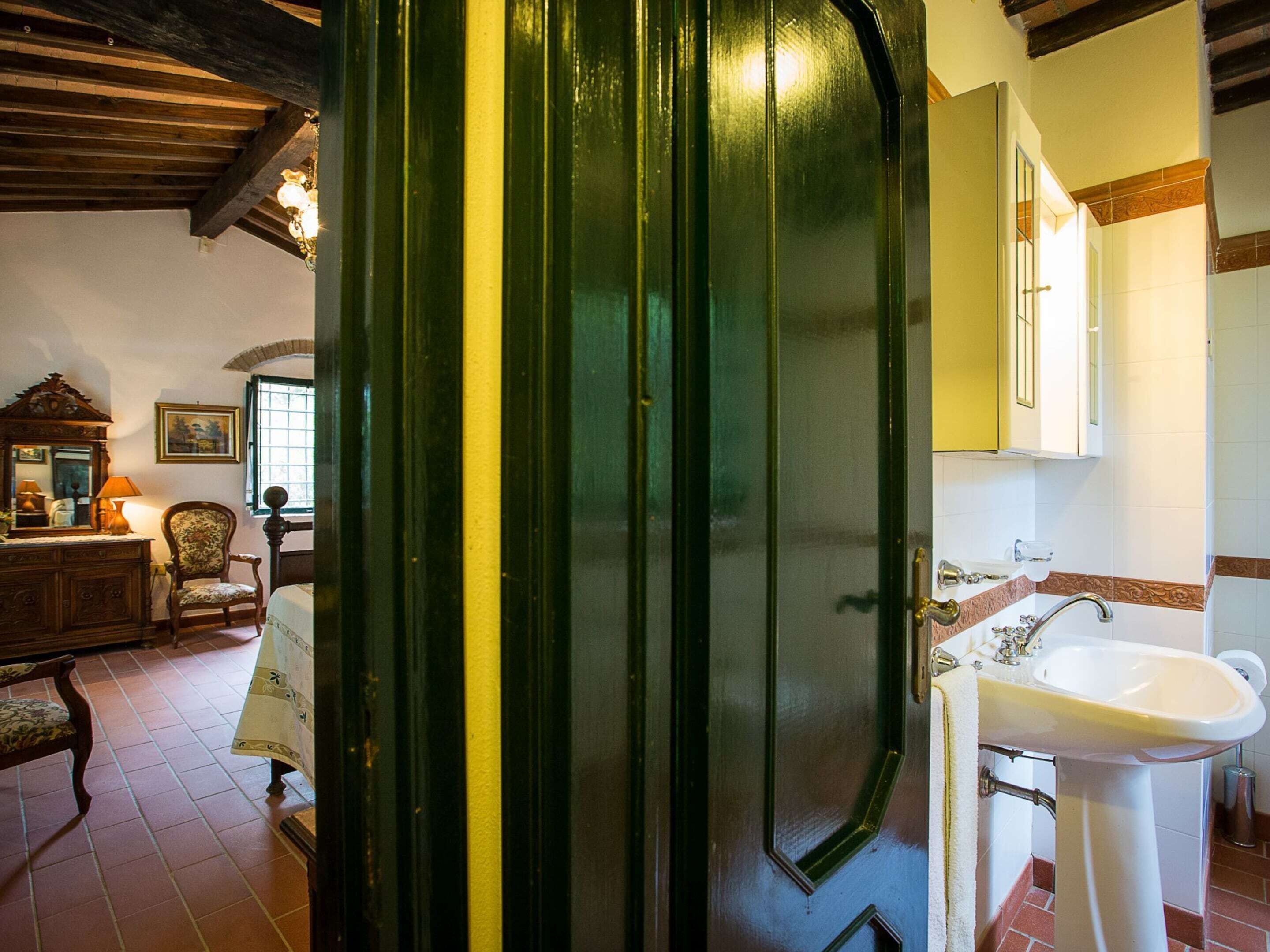 A room with a wooden ceiling, a green door, a bed, a dresser, a chair, a window, and a bathroom with a sink and mirror.