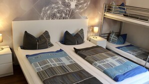 Quarto de hotel com duas camas, cada uma com roupa de cama e almofadas estampadas. Há uma cama beliche no canto e uma parede com luminária geométrica.