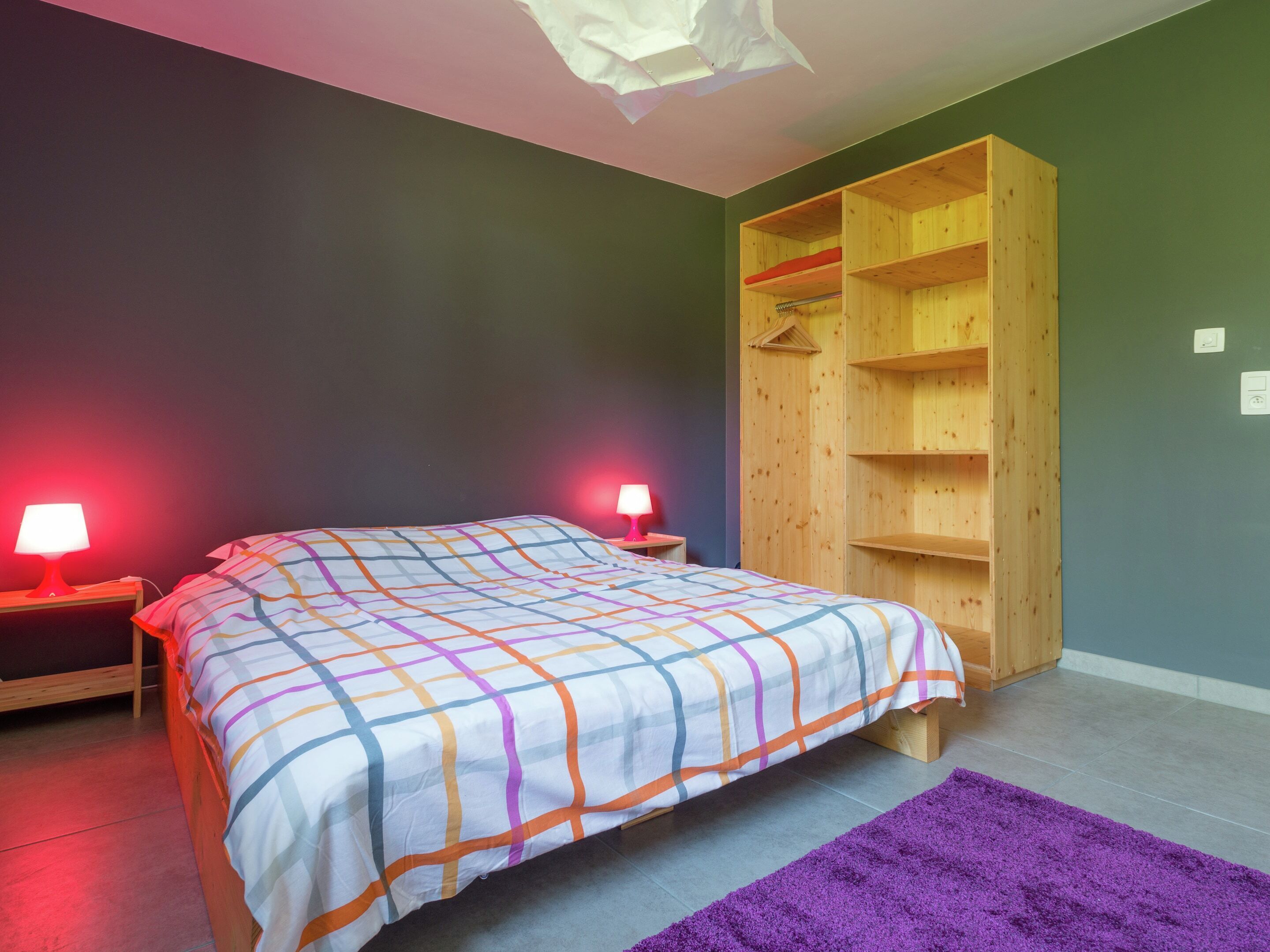 A bedroom with a bed, two bedside lamps, a purple rug, and a wooden wardrobe.