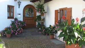 Une cour pavée, avec une porte en bois, des plantes en pot et un bâtiment blanc aux volets en bois.