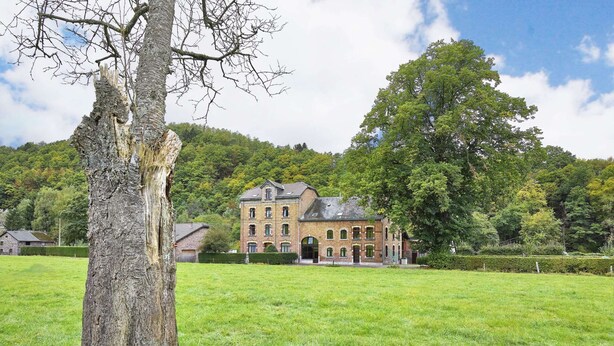 A large, historic brick building with multiple windows and a prominent entrance, surrounded by lush green fields and trees.