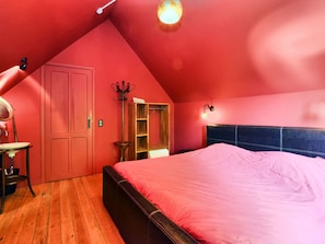 A bedroom with a sloped ceiling, a bed with a pink bedspread, a wooden wardrobe, and a small table with a chair. - A Beautiful Art Nouveau House With an Enormous Garden (Flobecq)