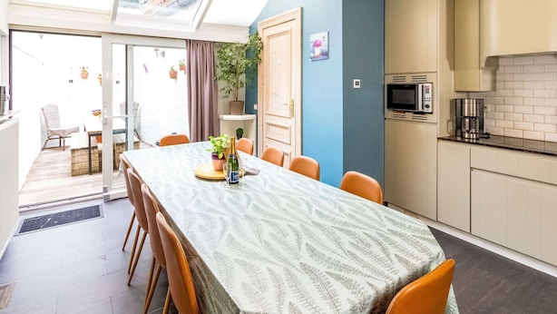 Ett modernt kök med inbyggd mikrovågsugn och kaffemaskin, en matplats med ett bord för fyra personer och en skjutdörr i glas som leder ut till en balkong med utemöbler.
