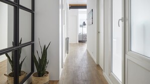 Un couloir doté d’un plancher en bois, de murs blancs et de miroirs à cadre noir. On y trouve deux paniers en osier contenant des plantes et une porte blanche au fond.