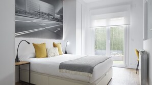 Une chambre moderne avec un grand lit, une table de chevet, un bureau et une fenêtre donnant sur un immeuble.