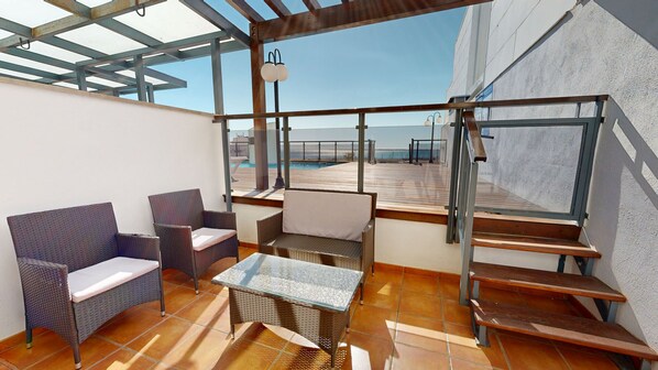 A sunlit patio with wicker furniture, a glass table, and a view of a pool and beach.