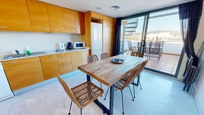 A modern kitchen with wooden cabinets, a dining table with wicker chairs, and a view of the city through a large sliding glass door. - APTO NAUTICO (Ampolla)