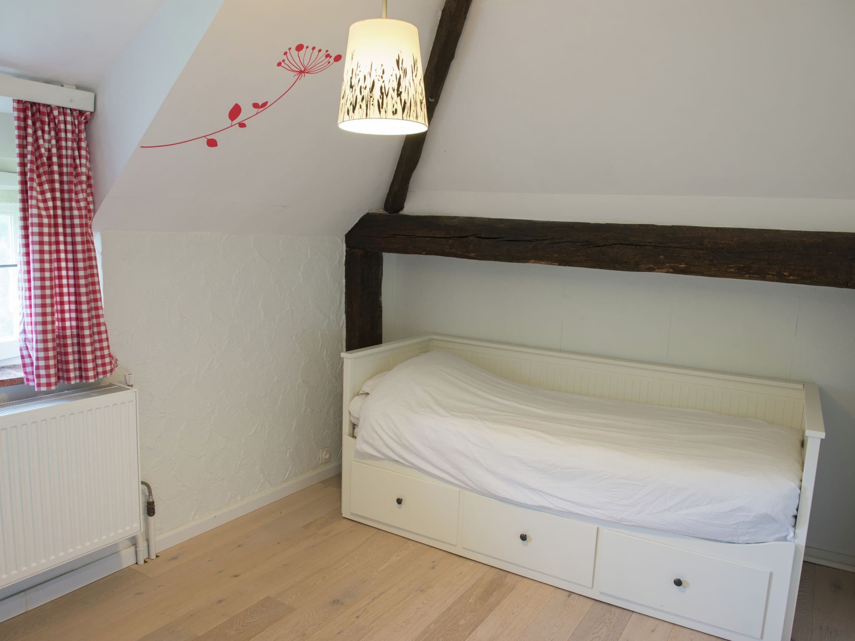 A bedroom with a single bed, a radiator, a window with checkered curtains, and a wall-mounted lamp.