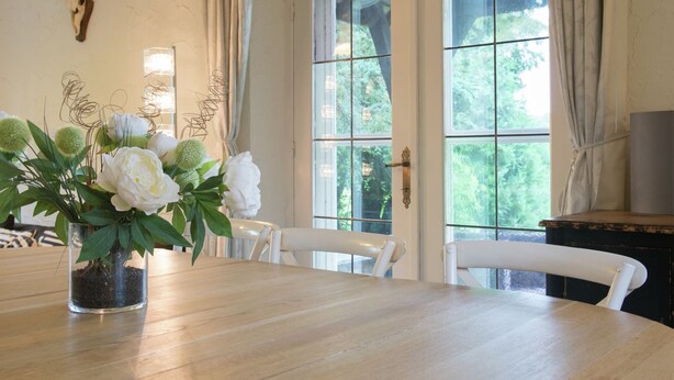 A dining area with a wooden table, white chairs, a vase with white flowers, and a deer skull on the wall. - Mansion in Spa Liege With Private Garden (Spa)
