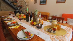 Mesa de jantar posta com pratos, talheres e diversos alimentos, incluindo frutas e uma garrafa de vinho, em um quarto com escada e um quadro na parede.
