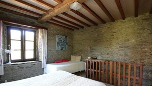 A bedroom with a stone wall, wooden beams, a bed, a nightstand, a lamp, and a window with curtains. - Forest Hideaway, Baillamont (Bievre)