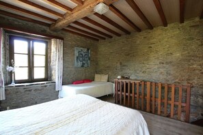 Een slaapkamer met een stenen muur, houten balken, een bed, een nachtkastje, een lamp en een raam met gordijnen.