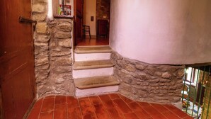 Un escalier aux murs en pierre et au sol recouvert de carreaux rouges mène à une pièce dotée d’une porte en bois et d’une petite étagère sur laquelle sont posés divers objets.