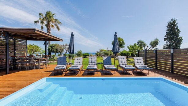 A swimming pool with lounge chairs and umbrellas, surrounded by a wooden deck and a view of the sea. - Nastazia Luxury Beach House (Ialisos)