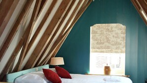 Un dormitorio con techo inclinado, cama con ropa de cama blanca y almohadas rojas, lámpara de noche y ventana con vista a un edificio.