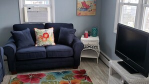 Un salon avec un canapé bleu foncé, un tapis à motifs floraux aux couleurs vives, un meuble TV en osier blanc et un tableau représentant une scène de plage.