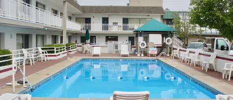 A swimming pool with lounge chairs and a covered area, surrounded by a building with multiple balconies and a parking area.