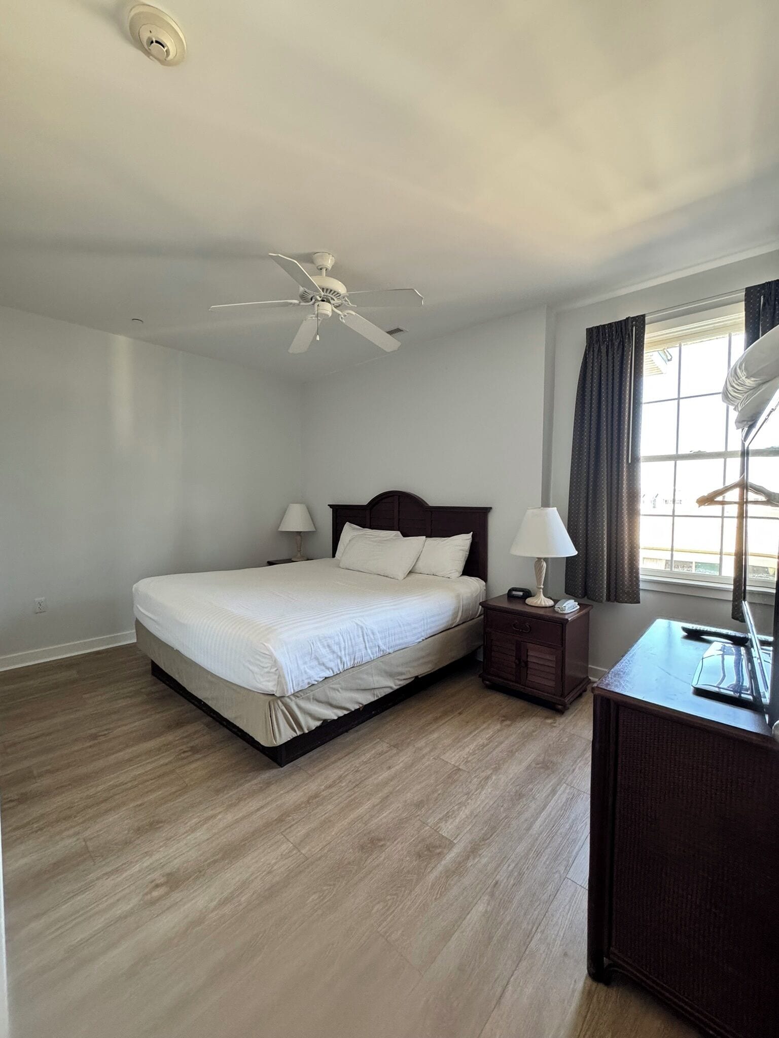 A bedroom with a bed, a nightstand, a desk, a ceiling fan, and a window with curtains.