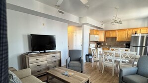 A modern living room with a sofa, coffee table, and TV. There is a dining area with chairs and a kitchen with cabinets and appliances. - Ebb Tide Suites (Ocean City)